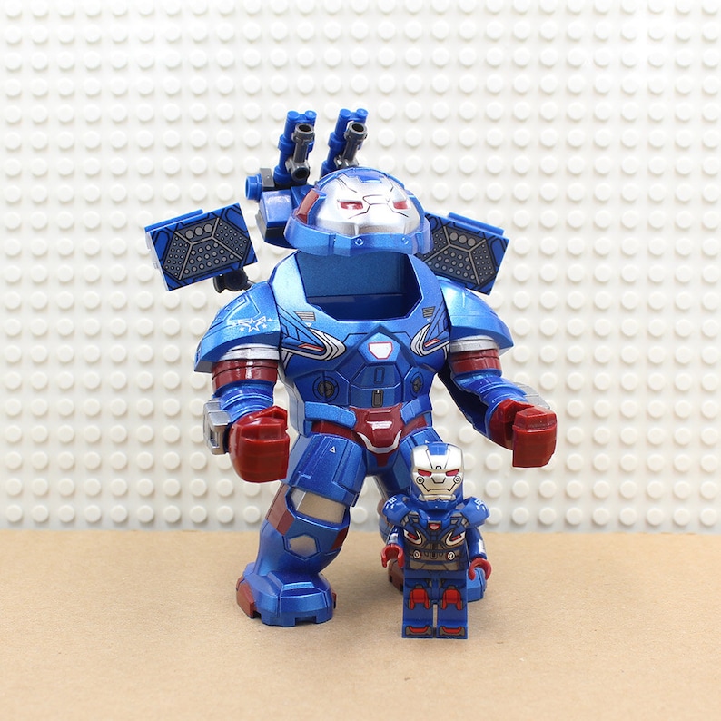 Może przedstawiać: Dwie niebieskie, czerwone i srebrne figurki zabawki superbohatera w robocim stroju. Większa figurka ma zamontowane na ramionach działa i szczeg&oacute;łowy design. Mniejsza figurka stoi z przodu, odzwierciedlając design większej.