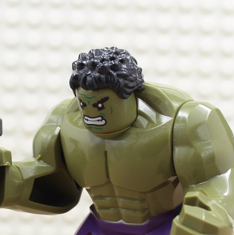 Puede incluir: Una minifigura Lego verde y negra del Hulk, con una construcci&oacute;n muscular y una expresi&oacute;n de enfado. La figura tiene una pieza de pelo negro y un torso verde con un pecho y un abdomen esculpidos.