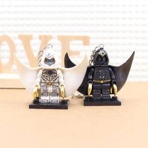 May include: Two minifigure keychains, one white and one black, with capes and gold accents. The white minifigure has a blue face and a crescent moon on its head. The black minifigure has a red face and a crescent moon on its head.