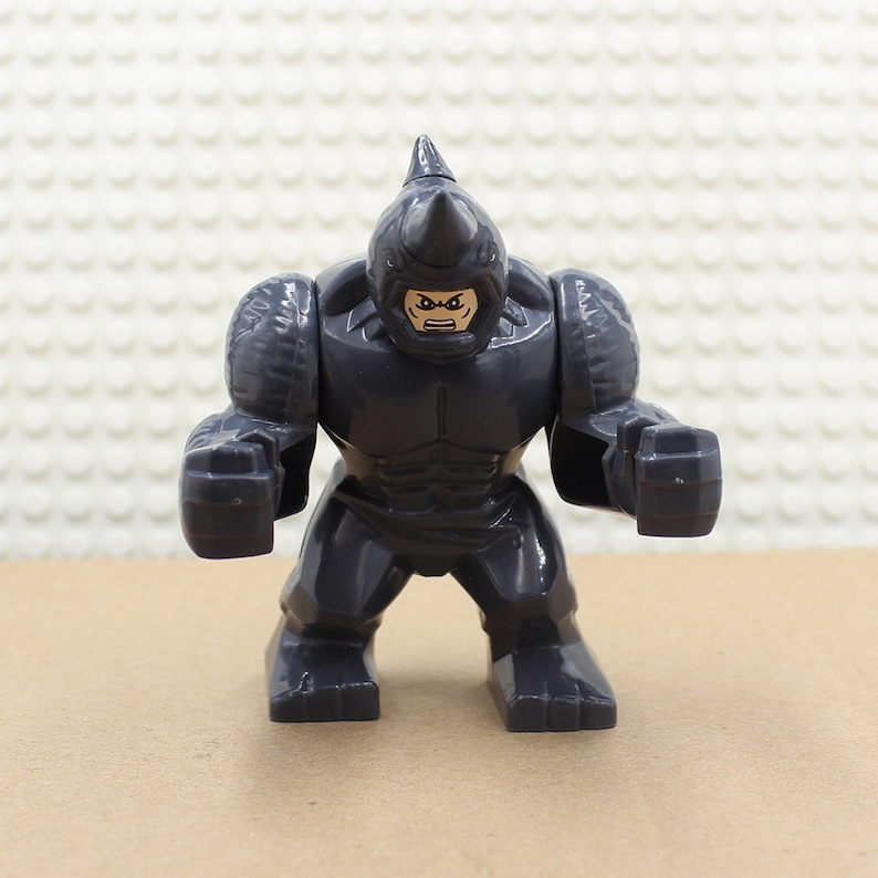 Puede incluir: Una minifigura Lego gris del personaje Rhino de Marvel Comics. La figura tiene una gran cabeza de rinoceronte gris con un cuerno negro y un cuerpo negro texturizado. La figura tiene una cara negra con una boca y ojos blancos.