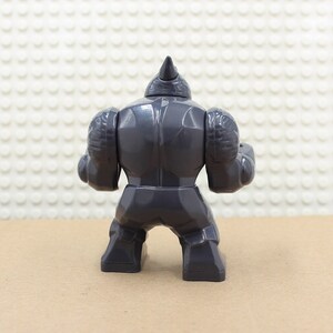 Puede incluir: Una figura de juguete de pl&aacute;stico gris de un rinoceronte, con un cuerno en la cabeza y una construcci&oacute;n muscular. La figura est&aacute; en posici&oacute;n de pie con los brazos a los lados.