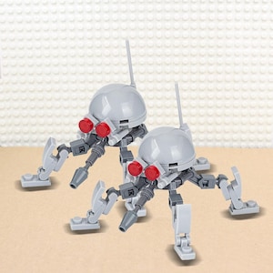 May include: Two gray and red toy robots with articulated legs and a dome-shaped head. The robots have a small antenna on top of the dome.