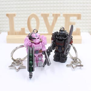 May include: Two pink and black Power Armor keychain figures with star-shaped keychains. The figures are holding weapons and are standing on a wooden surface with the word "LOVE" written on it.