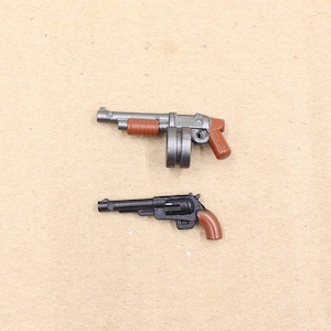 May include: Two toy guns: a gray and brown shotgun and a black and brown revolver. The shotgun has a cylindrical magazine and a brown grip. The revolver has a brown handle. Both are small and detailed.