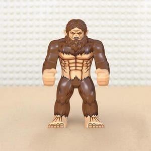 May include: A brown and tan action figure of a muscular humanoid beast with a furry chest and head. The figure has a stern expression and is posed with its arms at its sides. The figure's feet are large and detailed.