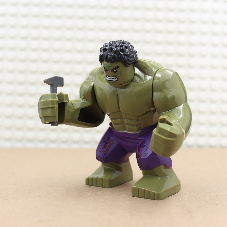 Puede incluir: Minifigura Lego Hulk verde con pelo negro, pantalones morados y un martillo gris en la mano.