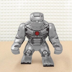 May include: A silver-colored Iron Man action figure with a red circular chest piece. The figure has a detailed design, including lines and dots, and is posed with its arms at its sides. The figure is made of plastic.
