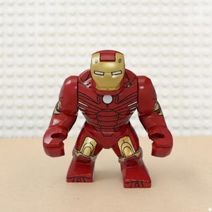 May include: A red and gold Iron Man action figure. The figure has a red body with gold accents on the helmet, shoulders, and legs. The figure is standing upright with its arms at its sides.