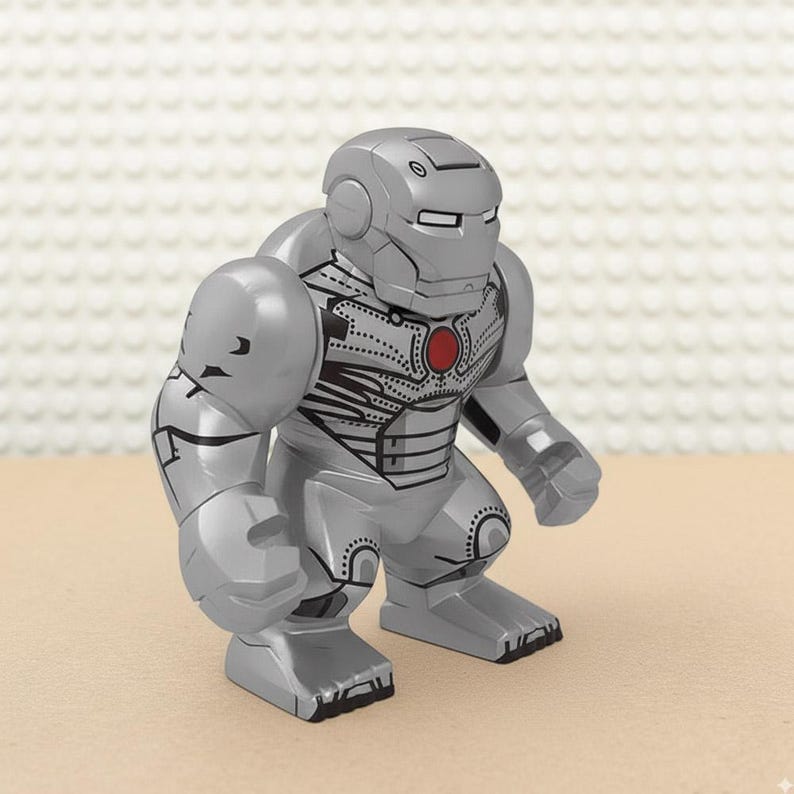 May include: A silver-colored toy figure resembling a superhero in a robotic suit. The figure has a helmet, broad shoulders, and a red circular emblem on its chest. Black detailing accents the suit's design.