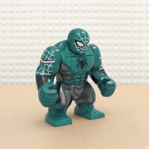 May include: A teal and gray action figure resembling a superhero. The figure has a spiderweb design on the head and arms, with a black spider emblem on the chest. The figure is in a standing pose.