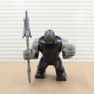 May include: A gray and black Lego minifigure of a character with a large head and a spear. The character has red eyes and a dark gray face. The spear is silver and has a sharp point.