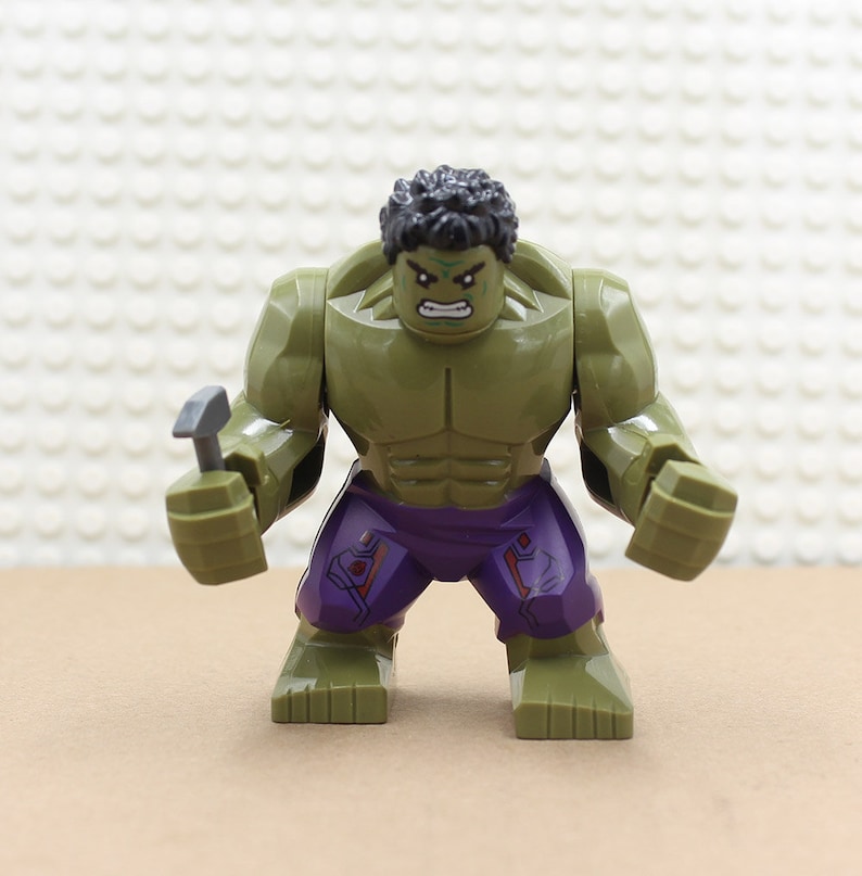 Puede incluir: Minifigura Lego Hulk verde con un torso y piernas con estampado morado y rojo. La minifigura sostiene un martillo plateado en su mano derecha.