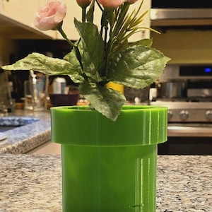 May include: A green 3D printed planter shaped like a Super Mario Bros. pipe with pink flowers inside.
