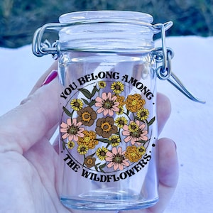 May include: A clear glass jar with a hinged lid and a metal clasp. The jar features a floral design with the text "YOU BELONG AMONG THE WILDFLOWERS." The flowers are in shades of pink, brown, and yellow.