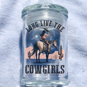 May include: Clear glass jar with a lid, featuring a graphic of a cowgirl on a horse in a desert landscape. The text "LONG LIVE THE COWGIRLS" is printed on the jar. The jar is designed to hold items.