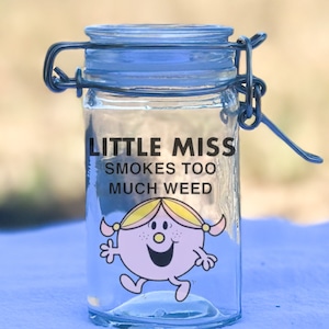 May include: A clear glass jar with a blue lid and metal clasp. The jar features the text "LITTLE MISS SMOKES TOO MUCH WEED" and an illustration of a cartoon character. The jar is designed for storage.