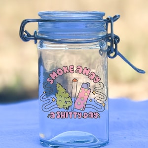 May include: A clear glass jar with a blue lid and metal clasp. The jar features a graphic with the text "Smoke Away a Shitty Day" and illustrations of a cannabis bud, a lit joint, and a lighter with smiley faces.
