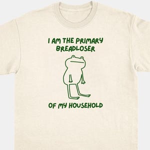 May include: A cream-colored short-sleeve t-shirt featuring a green line-art illustration of a frog centered on the chest. Above the frog, the text reads I AM THE PRIMARY BREADLOSER and below the frog, the text reads OF MY HOUSEHOLD in green.