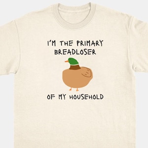 Puede incluir: Una camiseta de manga corta de color crema con un gráfico de un pato real en el centro. Sobre el pato, el texto dice I'M THE PRIMARY BREADLOSER, y debajo del pato, el texto dice OF MY HOUSEHOLD en una fuente negra sencilla.
