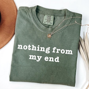 May include: Olive green t-shirt with the phrase "nothing from my end" in white block letters. A gold necklace with a small pendant rests on the shirt. A brown hat and dried palm leaves are in the background.
