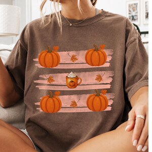 May include: A brown short-sleeve t-shirt featuring a graphic print of four orange pumpkins, scattered autumn leaves, and a central mug of coffee with whipped cream, arranged across three horizontal light pink brushstroke stripes.