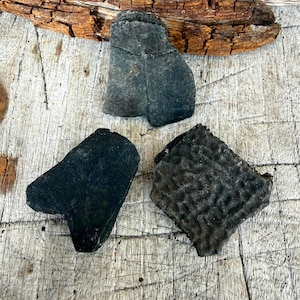May include: Three dark gray, irregularly shaped stones are arranged on a weathered wooden surface. The stones vary in texture, with one appearing smooth, another with a rough, porous surface, and the third with a slightly textured surface. A piece of driftwood is in the background.