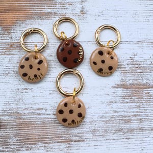 Puede incluir: Cuatro llaveros dorados con dijes en forma de galleta marrón y beige. Cada dije tiene lunares negros y una inscripción dorada: "Cookie", "Benji", "Barry" y "Willow".