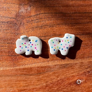 Op de afbeelding: Twee witte, platte, diervormige bedels met een kleurrijk stippenpatroon van kleine blauwe, roze, gele en groene stippen. Elke bedel heeft een klein gaatje bij de bovenkant, waardoor deze als hanger of decoratief element kan worden gebruikt.