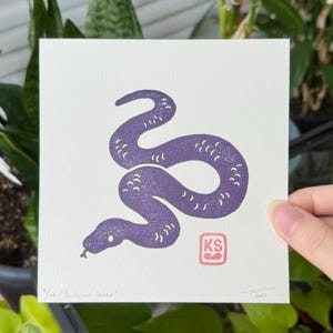 Puede incluir: Una impresión cuadrada con una ilustración de una serpiente morada. La serpiente está estilizada con líneas curvas y pequeños patrones de puntos. La impresión incluye el texto "9/ve 'Sunbeam Snake'" y un cuadrado rojo con las letras "KS".