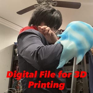 Op de afbeelding: Een blauw en wit 3D-geprint masker met de rode tekst "Digital File for 3D Printing". Het masker heeft een vloeiende, organische vorm. Een persoon houdt het masker vast.