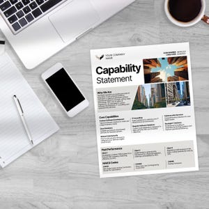 May include: A white Capability Statement document with the company name and logo, a smartphone, a laptop, a pen, and a cup of coffee on a light wood-grain surface. The document lists core capabilities and past performance.