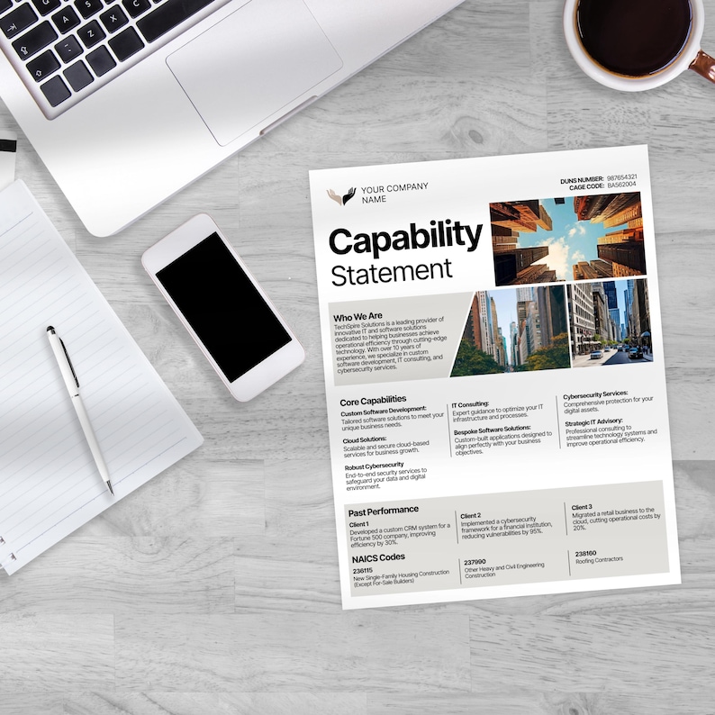May include: A white Capability Statement document with the company name and logo, a smartphone, a laptop, a pen, and a cup of coffee on a light wood-grain surface. The document lists core capabilities and past performance.