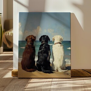 May include: A canvas print depicts three dogs of different colors, brown, black, and white, sitting side-by-side on a sandy beach, facing the ocean. The dogs are wearing collars. The background shows a blue sky with clouds and the ocean.