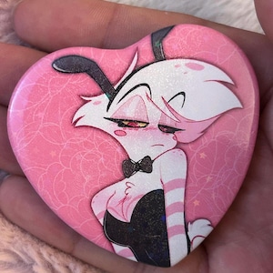 May include: A heart-shaped button featuring a cartoon character with white hair, black bunny ears, and a black bow tie. The character is wearing a black outfit with pink and white striped sleeves, set against a pink background with a web-like pattern.