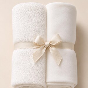 May include: Two folded white blankets secured together with a light beige ribbon tied in a bow. One blanket features a textured, nubby surface, while the other blanket has a smooth, soft fleece-like texture. The items are presented as a set.