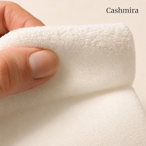 May include: A soft, off-white fabric with a dense, fuzzy texture being held. The word Cashmira is printed in black in the upper right corner. The material appears thick and plush, showing a folded edge that highlights the pile of the textile.