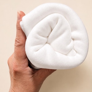 May include: A soft, solid white fabric item folded into a compact, rounded shape. The material has a smooth, plush texture and a clean, uniform appearance, showing a dense weave that creates gentle folds and shadows within the rolled structure.