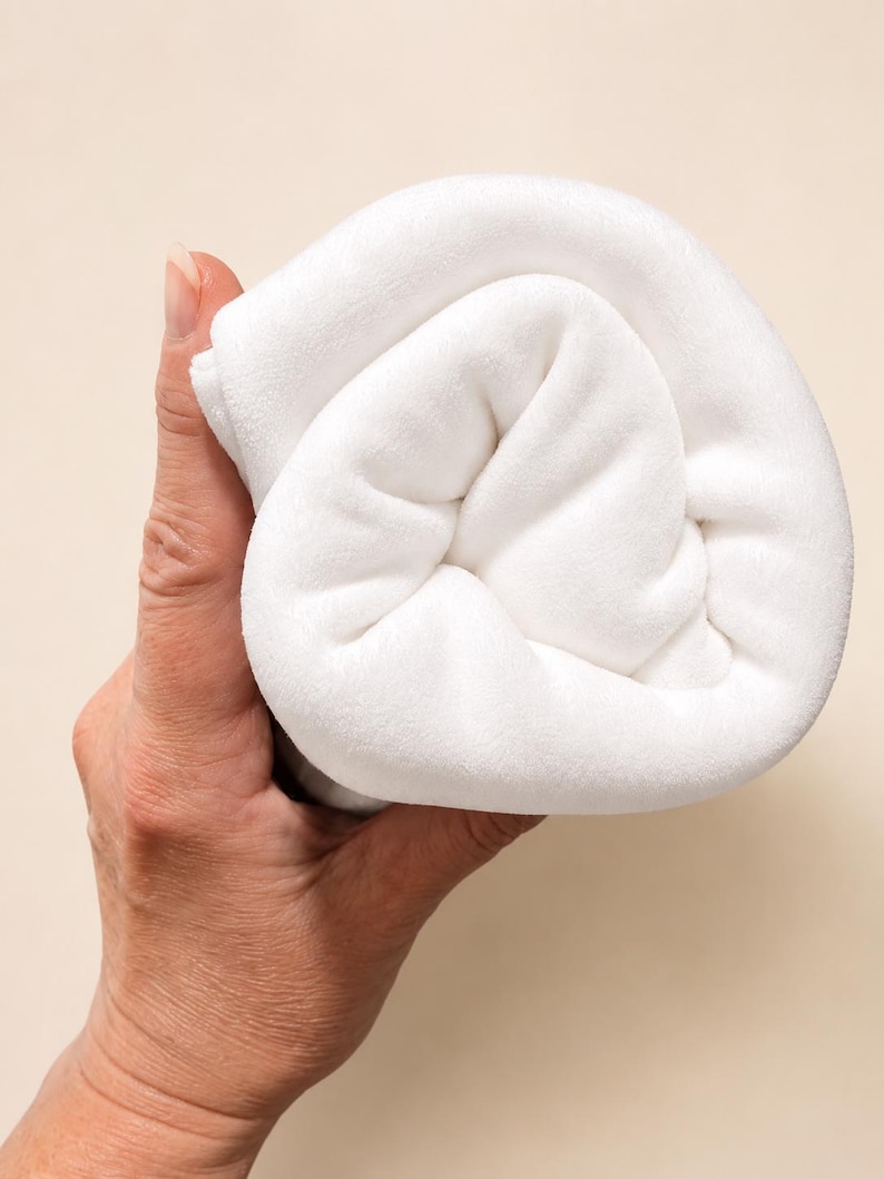 May include: A soft, solid white fabric item folded into a compact, rounded shape. The material has a smooth, plush texture and a clean, uniform appearance, showing a dense weave that creates gentle folds and shadows within the rolled structure.