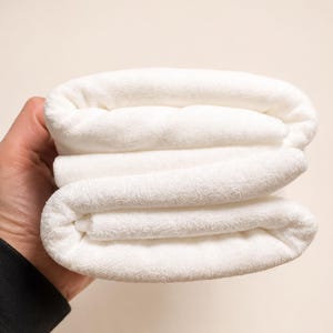 May include: A folded white towel with a soft, plush, and textured surface. The fabric appears thick and absorbent, featuring a consistent, clean white color throughout the entire piece. The towel is neatly layered in multiple horizontal folds.