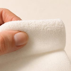May include: A piece of white fabric with a soft, plush, and fuzzy texture. The material is folded over, revealing a thick, dense pile surface that appears uniform in colour and texture throughout the visible portion of the textile material.