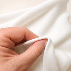May include: A piece of solid white fabric with a soft, matte, and slightly fuzzy texture. The material is folded and draped, showing a consistent opaque white color throughout the surface. The fabric appears to be a thick, plush textile.