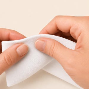 May include: A rectangular piece of white fabric with a fine, textured surface. The material appears soft and pliable, held between fingers to demonstrate its flexibility and matte finish. The fabric is opaque, clean, and uniform in color.