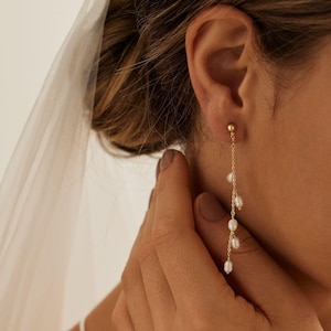 May include: Gold dangle earrings with small white pearls hanging from a delicate chain. The earrings are worn by a person with a veil in their hair.