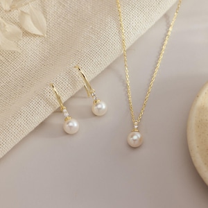 Puede incluir: Un collar de oro y un par de pendientes a juego, cada uno con una perla blanca suspendida de un pequeño engaste dorado acentuado con una diminuta piedra clara. El conjunto de joyas se muestra con el texto DaintyMinimalistShop.