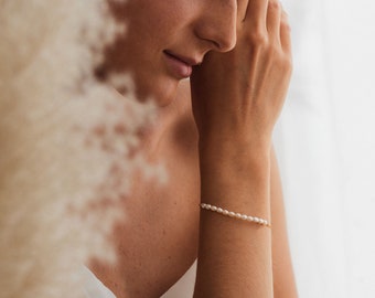 May include: A delicate bracelet featuring a single strand of small, white, oval-shaped freshwater pearls strung on a thin gold-toned chain. The bracelet is worn around the wrist, showcasing the uniform size and smooth texture of the pearls.