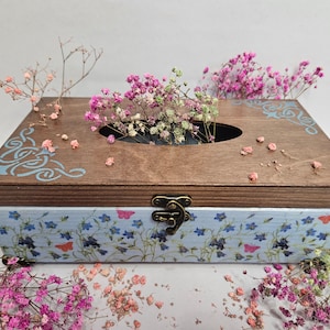 Könnte beinhalten: Ein hölzerner Taschentuchbox-Deckel mit einem blauen Blumendesign und einem hellbraunen Deckel. Der Deckel hat ein blaues und weißes Wirbeldesign und eine kleine Öffnung für Taschentücher. Die Box ist mit getrockneten Blumen verziert.