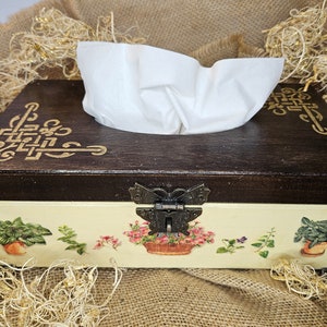 Könnte beinhalten: Ein hölzerner Taschentuchbox-Deckel mit einem dekorativen Blumendesign und einem Schnappverschluss in Form eines Schmetterlings. Die Box ist in Cremefarben mit braunen Akzenten lackiert und hat ein goldfarbenes Design.