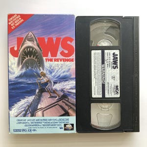 May include: A Jaws The Revenge VHS tape and its cardboard sleeve. The sleeve features a shark emerging from the ocean behind a person on a boat, with the text "Contains new footage not seen in U.S. theaters" and "This Time It's Personal."