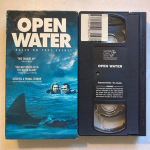 May include: A VHS tape and its case for the movie "Open Water." The case is blue with the title in large white letters and a shark fin graphic. The tape is black with the title and Lions Gate Entertainment logo.