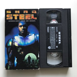 Peut inclure: Une cassette VHS du film "Steel". La face avant de l'étui présente un homme noir en costume futuriste, avec le titre "Steel" et le slogan "Heroes don't come any bigger." Le dos de l'étui porte le titre du film et la classification.